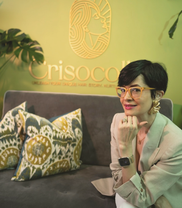 Cindy Suârez, founder of crisocolla sittingon a sofa . background is a green wall with Crisocolla logo design placed on top of the sofa. Cindy is wearing a crewmy beige blazer and is smiling sideway. 