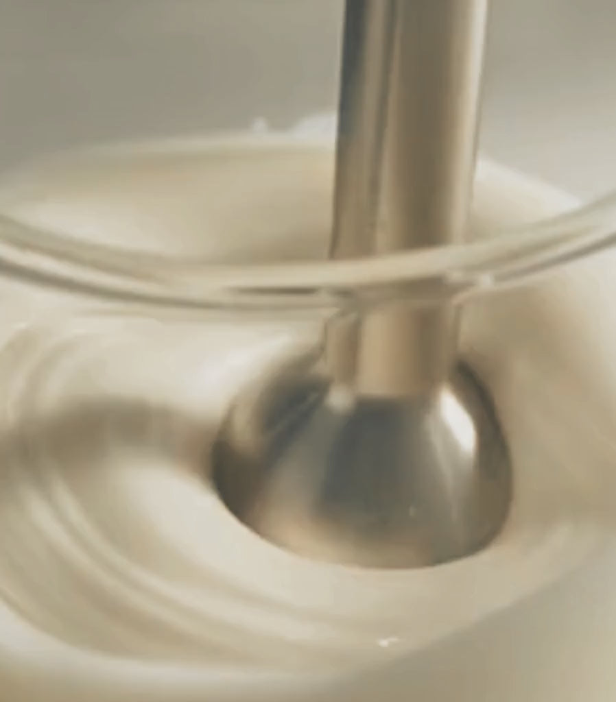 A cinematic close-up of the Crisocolla alchemical process: a laboratory blender mixing the creamy BotaniReset Hair Balm formula inside a glass beaker, set against a minimalist white background and clean white laboratory tiles.